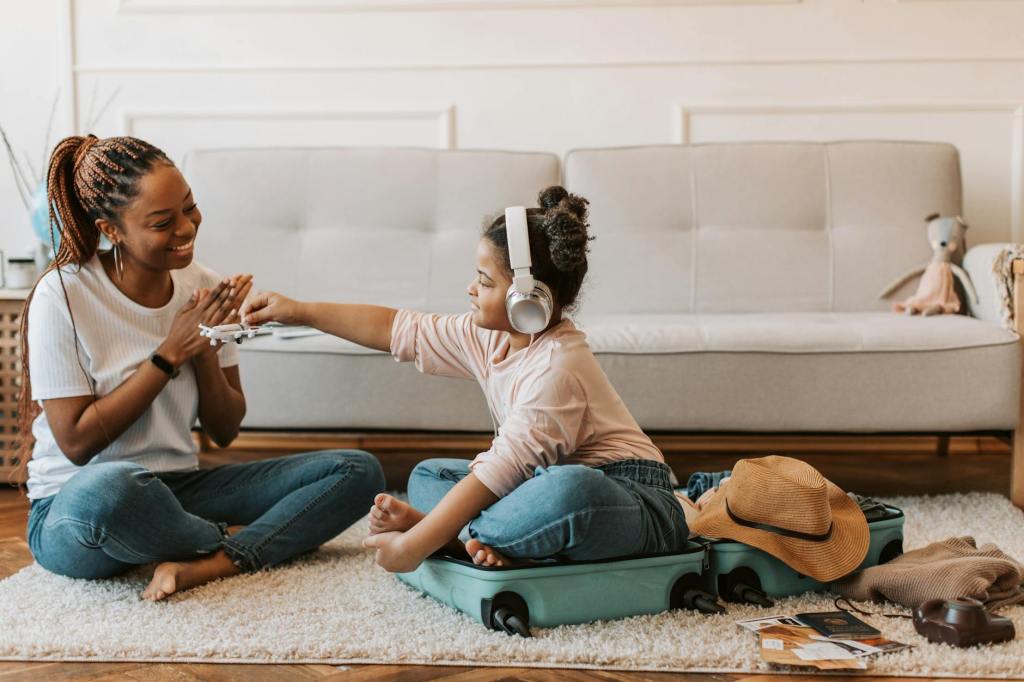 A mother and child playing together at home. The child is sitting in a suitcase wearing headphones, while the mother sits on the floor nearby. They are reaching out and touching hands, sharing a playful and connected moment.